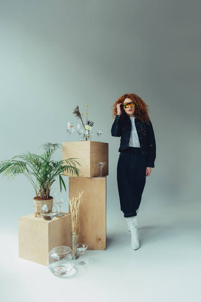 fashionable redhead girl posing near wooden boxes, glasses and plants on grey - Photo, image