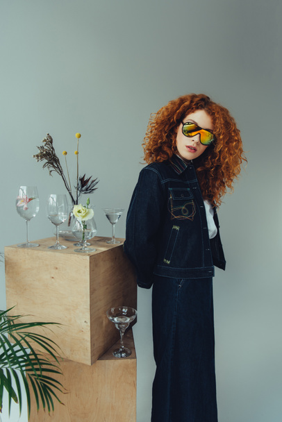 fashionable redhead girl in sunglasses posing near wooden boxes, glasses and plants on grey - Photo, Image