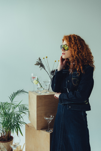 trendy redhead girl posing near wooden boxes, glasses and plants isolated on grey - Zdjęcie, obraz