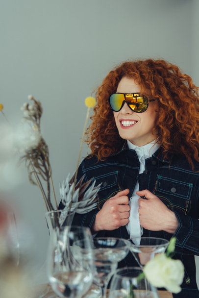 smiling stylish redhead woman in sunglasses posing near glasses and dried flowers isolated on grey - Zdjęcie, obraz