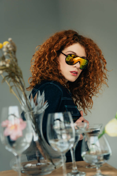 stylish redhead woman in sunglasses posing near glasses and dried flowers on grey - 写真・画像