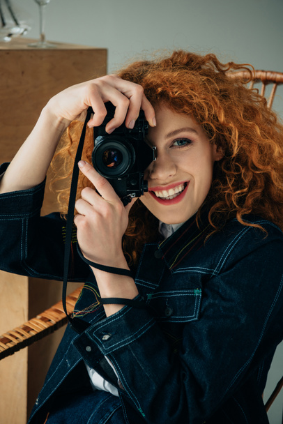 灰色で隔離されたフィルムカメラで写真を撮るウィッカーチェアで笑顔のスタイリッシュな赤毛の女性 - 写真・画像