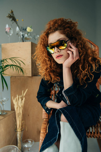 stylish redhead girl in sunglasses in wicker chair near wooden boxes, glasses and plants on grey - Photo, Image