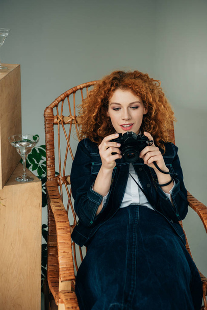 beautiful stylish redhead woman in wicker chair taking photo with film camera on grey - Foto, Imagen
