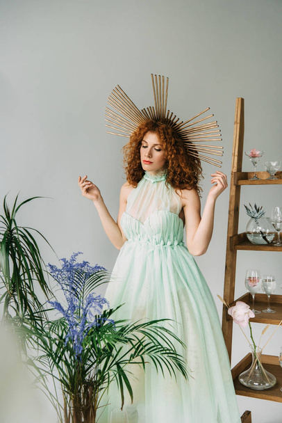 redhead girl with accessory on head in dress posing near plants and ladder - Photo, image