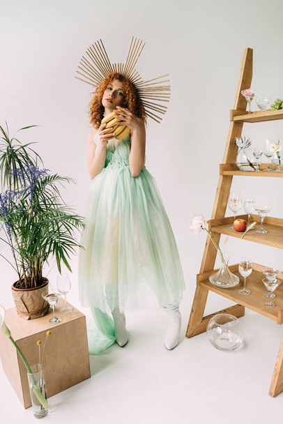 stylish redhead girl with accessory on head and bananas near ladder with flowers and glasses - Photo, Image