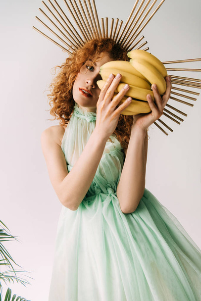 redhead woman with accessory on head posing with bananas on white - Zdjęcie, obraz