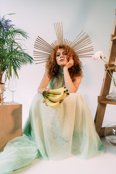 stylish redhead woman with accessory on head posing with bananas and propping head - Zdjęcie, obraz