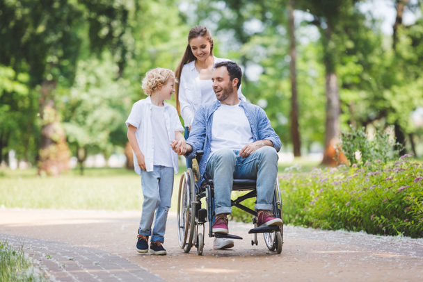 красивая мама катает коляску с отцом-инвалидом в парке рядом с сыном
 - Фото, изображение