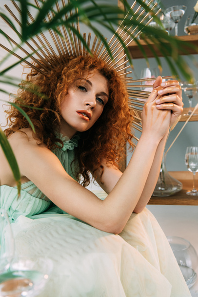 fashionable redhead girl with accessory on head in dress posing near plant - Foto, imagen