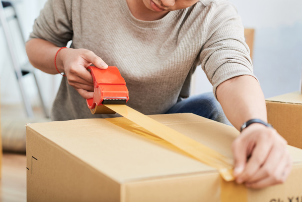 Unrecognizable young man using sticky tape to seal carton box while relocating into new flat - Photo, Image