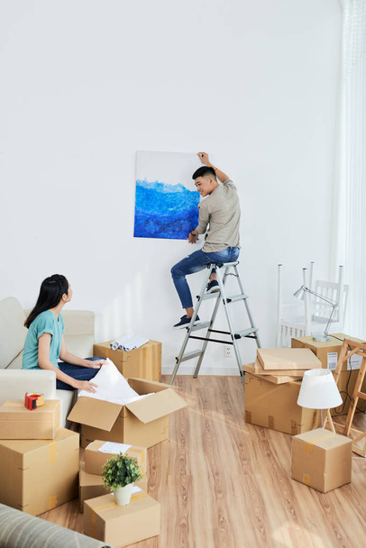Young Asian man sitting on ladder and asking girlfriend where to hang picture while relocating into new flat - Photo, Image