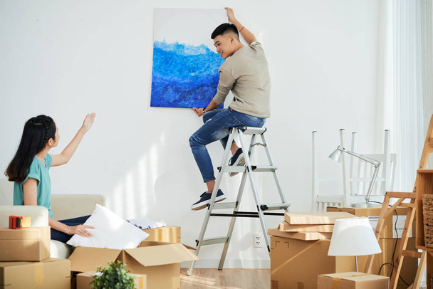 Young Asian lady showing boyfriend where to hang nice picture while decorating new apartment together - Zdjęcie, obraz
