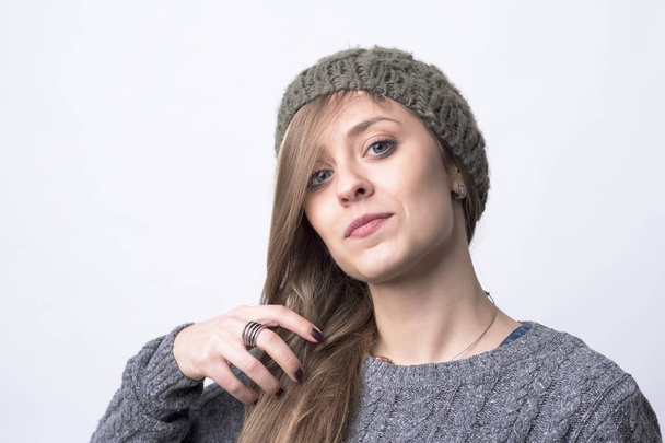 Confident funky hipster girl with knitted gray cap brushing hair looking at camera on light gray background.  - Photo, Image