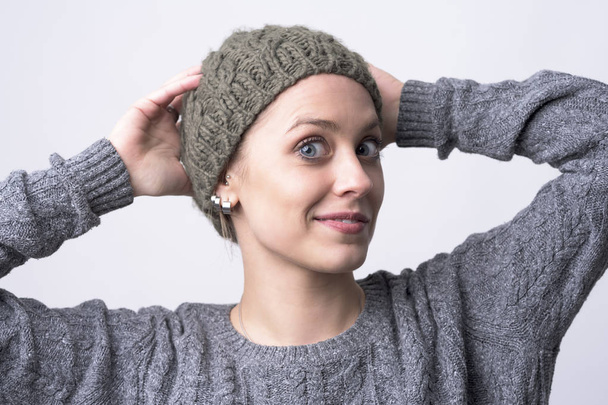 Cute funny blue eyes young woman trying on gray knitted cap on light gray background.  - Фото, зображення
