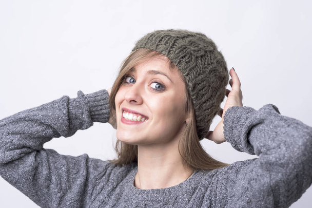 Cute charming young woman trying on gray knitted beanie cap smiling at camera on light gray background.  - Photo, image