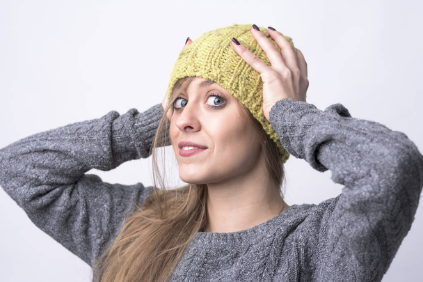 Retrato de una linda joven confiada probándose la gorra de punto amarillo para clima frío de invierno sobre fondo gris claro
.  - Foto, Imagen