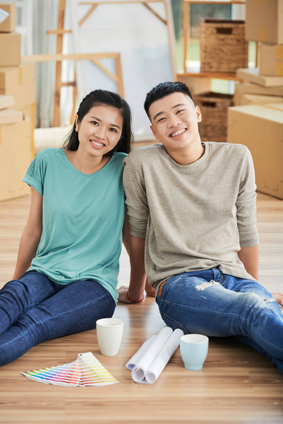 Ευτυχισμένο νεαρό ασιατικό ζευγάρι κάθεται στο νέο τους σπίτι με τα σχέδια δίπλα τους - Φωτογραφία, εικόνα