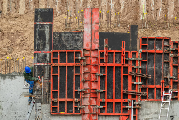 Будівельник, який працює на будівельному фундаменті. Чоловік у синьому робочому одязі на драбині пасує до металу для зміцнення бетонної стіни на схилі пагорба
 - Фото, зображення