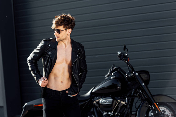 黒いオートバイの近くに立っている革のジャケットで筋肉の胴体を持つ若い男 - 写真・画像