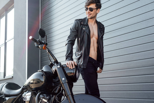  young man in sunglasses with leather jacket and naked torso standing near black motorcycle, smiling and looking away  - Fotó, kép