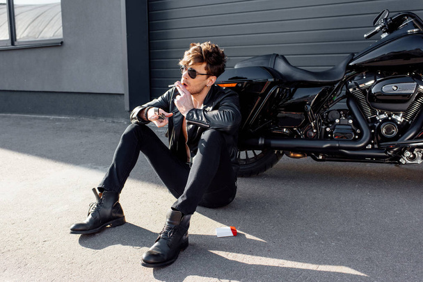 handsome young man with cigarette and sunglasses sitting on ground near motorcycle - Fotoğraf, Görsel