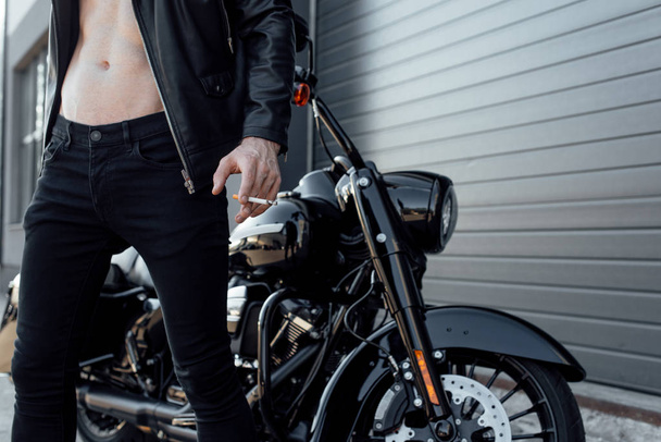 vista recortada del hombre con el torso desnudo sosteniendo cigarrillo y de pie cerca de la motocicleta
 - Foto, imagen