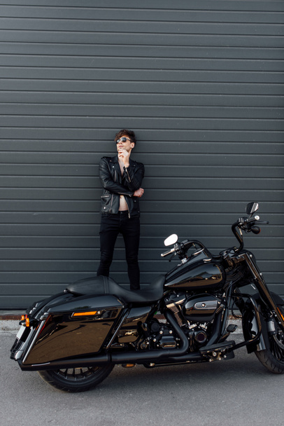 full length view of young man smoking cigarette while leaning on wall and standing near motorcycle - Photo, Image