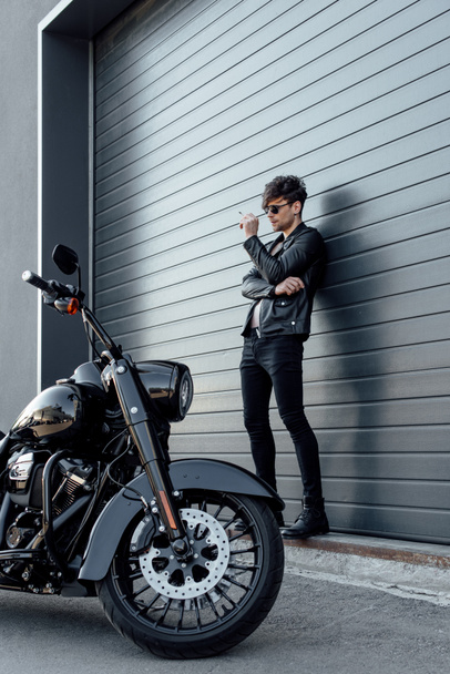 full length view of handsome motorcyclist in sunglasses standing near wall close to motorcycle and holding cigarette  - Foto, immagini