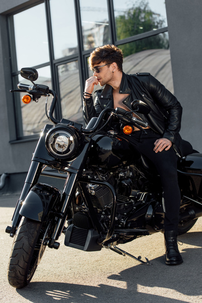 Красивый мужчина в солнечных очках в черной одежде сидит на мотоцикле и смотрит в сторону
 - Фото, изображение