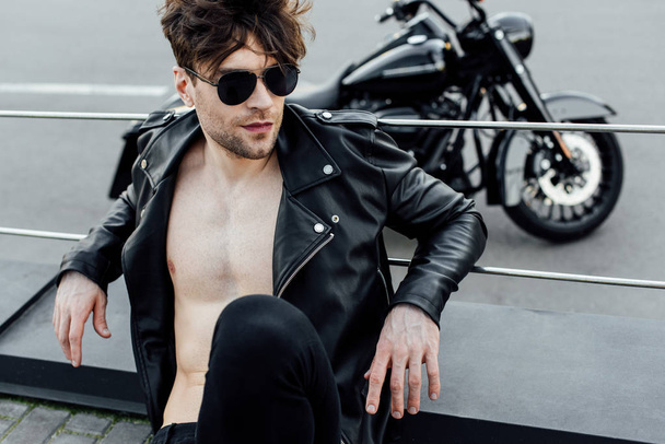selective focus of man with naked torso in leather jacket sitting on ground with motorcycle on background - Фото, изображение