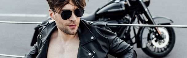 panoramic shot of man with naked torso in leather jacket sitting on ground with motorcycle on background - Photo, Image
