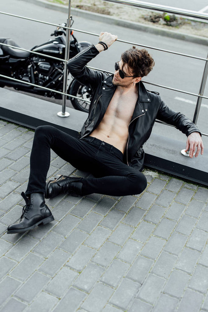 sexy motorcyclist in leather jacket with naked torso sitting on ground and holding metal fence - Φωτογραφία, εικόνα
