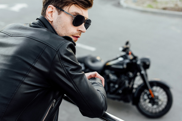 selective focus of young man in black leather jacket standing near metal fence with motorcycle on background - Фото, изображение
