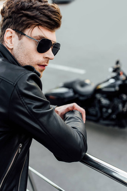 selective focus of young man in black leather jacket and sunglasses standing near metal fence with motorcycle on background - Fotó, kép