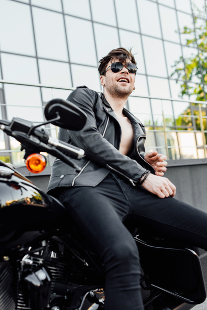 мотоциклист в солнцезащитных очках и черной кожаной куртке сидит на мотоцикле, улыбаясь и отводя взгляд
 - Фото, изображение