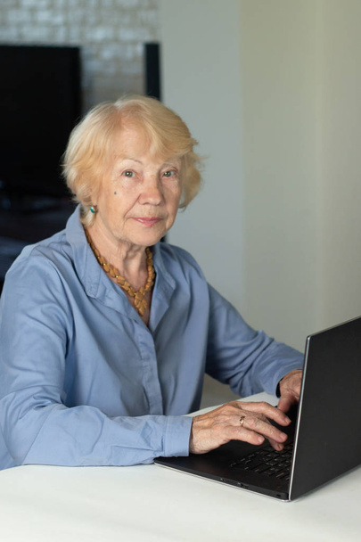 Ηλικιωμένη γκριζομάλλη γυναίκα με φορητό υπολογιστή. Ηλικιωμένη γυναίκα γράφοντας απομνημονεύματα σε φορητό υπολογιστή, αναζητώντας πληροφορίες στο διαδίκτυο ή επικοινωνώντας διαδικτυακά - Φωτογραφία, εικόνα