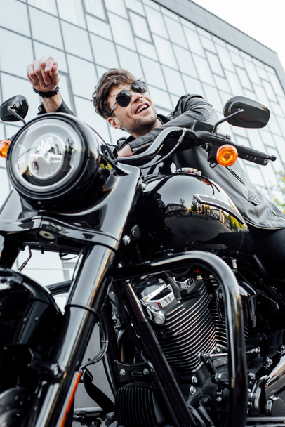 низкий угол зрения красивый мужчина в кожаной куртке сидит на мотоцикле, amiling и глядя в сторону
 - Фото, изображение
