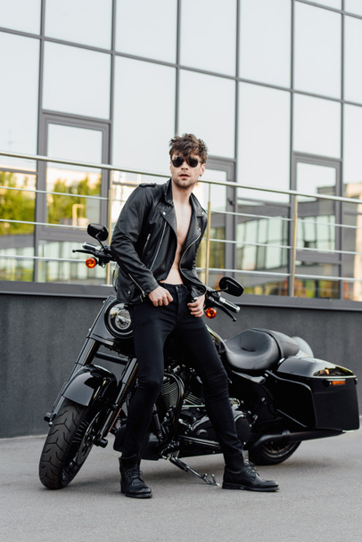 полный вид на мотоциклиста без рубашки, стоящего рядом с мотоциклом снаружи и смотрящего в сторону
 - Фото, изображение