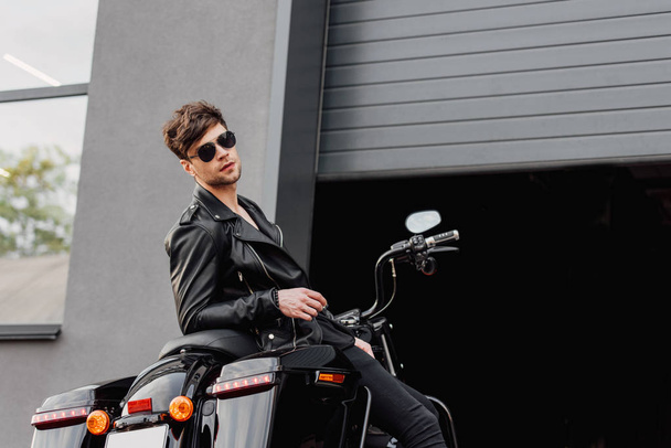 motorcyclist in sunglasses and leather jacket sitting on motorcycle near opened garage and looking at camera  - Foto, Bild
