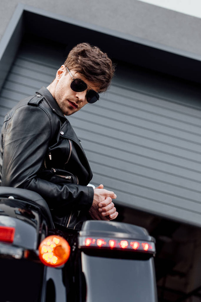 stijlvolle jonge man in zonnebril leunend op motorfiets en kijken naar camera - Foto, afbeelding
