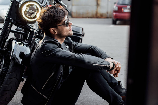 side view of handsome motorcyclist sitting on ground near motorcycle with lamp lighting up  - Foto, imagen