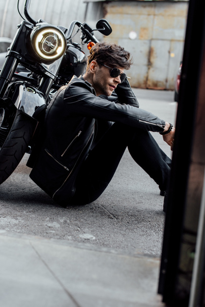 handsome young motorcyclist sitting on ground near black motorcycle with luminous lamp - Photo, Image