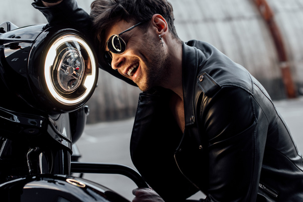 pohledný mladý muž, který se opírá o motocykl a usmíval se při pohledu na lampu  - Fotografie, Obrázek