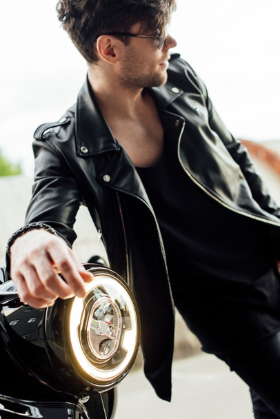 επιλεκτική εστίαση της φωτεινής λάμπας του κύκλου και όμορφος άνθρωπος σε γυαλιά ηλίου στέκεται κοντά μοτοσικλέτα - Φωτογραφία, εικόνα