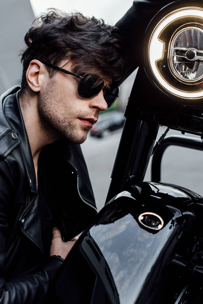 przystojny młody człowiek siedzi w pobliżu czarnego motocykla i pochylony głowę na kierownicy - Zdjęcie, obraz