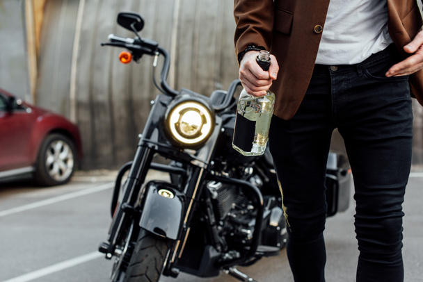 cropped view of man in jacket walking off motorcycle and holding bottle of alcohol  - Foto, Imagem