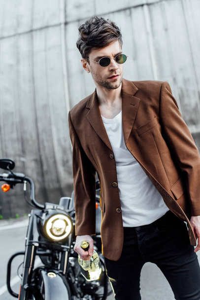 Selektiver Fokus eines gutaussehenden Mannes mit Sonnenbrille und brauner Jacke, der mit Alkoholflasche in Motorradnähe unterwegs ist  - Foto, Bild