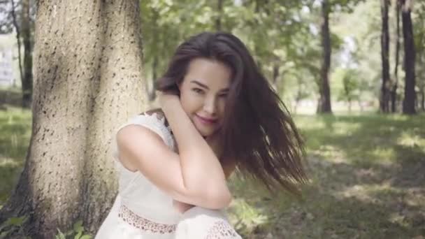 Portrait adorable young girl with long brunette hair wearing a long white summer fashion dress sitting under a tree in the park. Leisure of a pretty woman looking at the camera outdoors. - Video