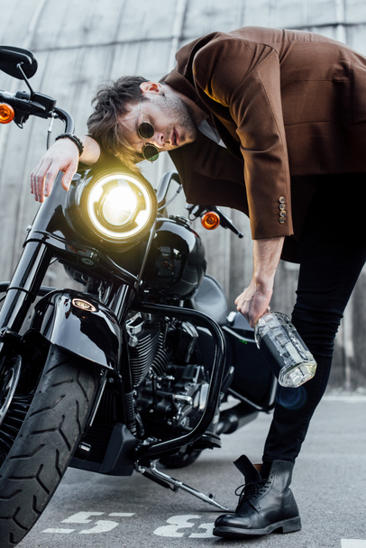 joven cabeza apoyada en el manillar de la motocicleta con lámpara luminosa mientras sostiene la botella con alcohol
 - Foto, Imagen